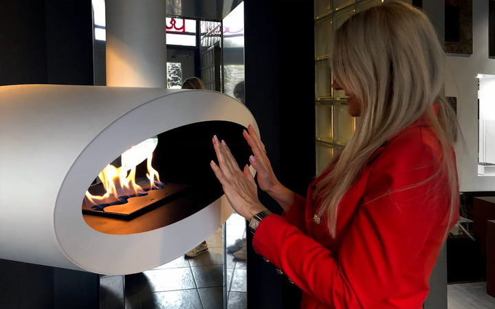 bioethanol-haengekamin-tube-2side-xl-weiss-ethanolkamin-decke-mit-flammen-vor-spiegel-in-wohnzimmer-blonde-frau-in-roten-mantel-waermt-haende-an-flammen