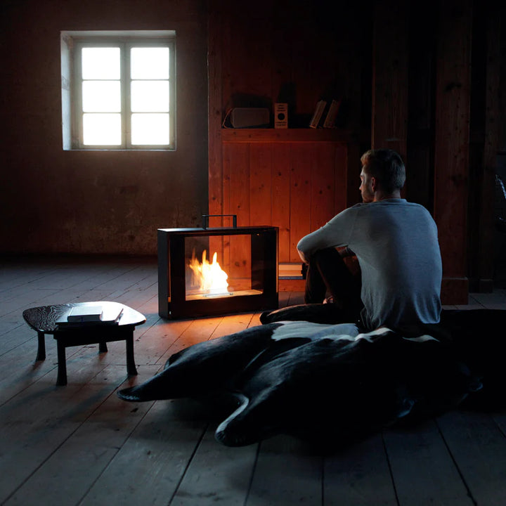 Bioethanol Standkamin TRAVELMATE von conmoto mit Flammen auf einem Holzboden, davor ein sitzender Mann